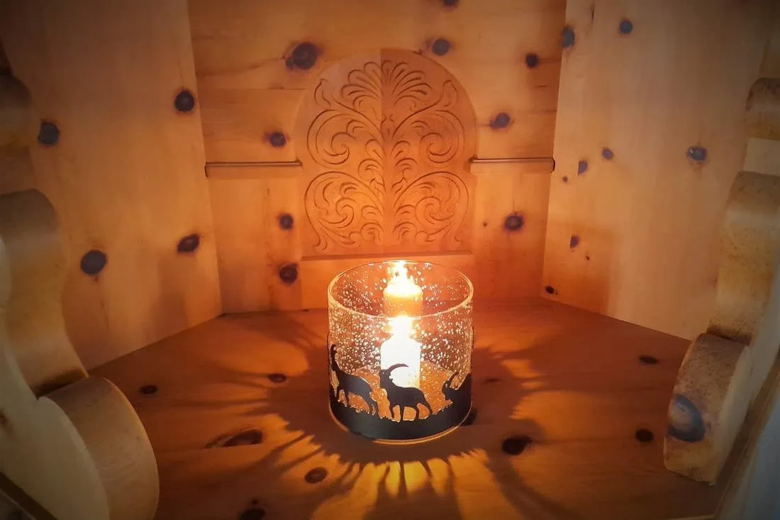 Windlicht mit brennender Kerze in einer Nische aus Arvenholz, dahinter ein geschnitztes Ornament.