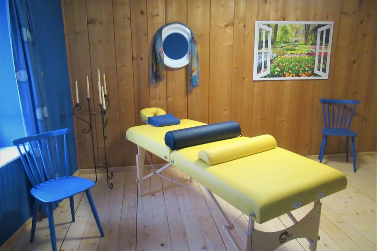 Behandlungsraum mit gelber Massageliege, zwei blauen Stühlen und Wänden aus Holz.