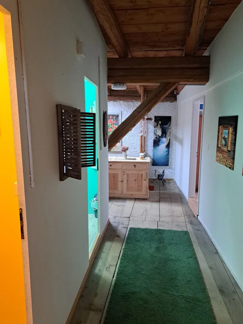 Gang mit Steinplatten und grünem Läufer, Holzbalken an der Decke und Bildern an den Wänden.