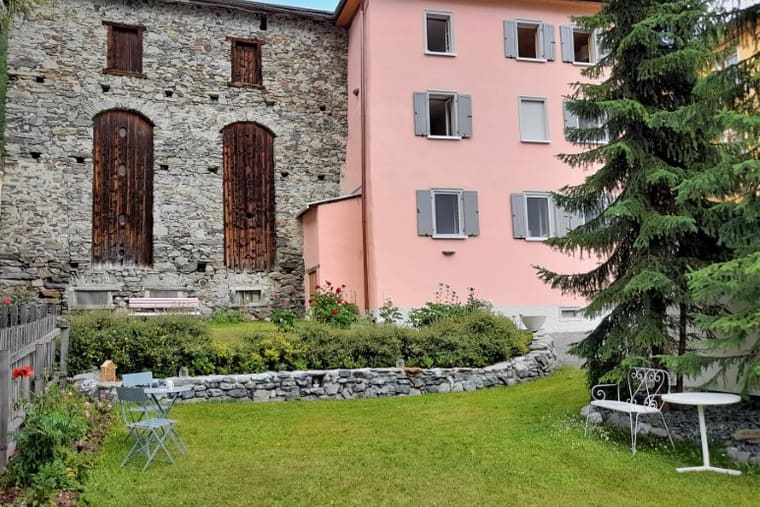 Das rosa verputzte Haus mit grüngrauen Fensterläden neben dem Steinstall mit zwei hohen Holztoren, davor der Rasen.
