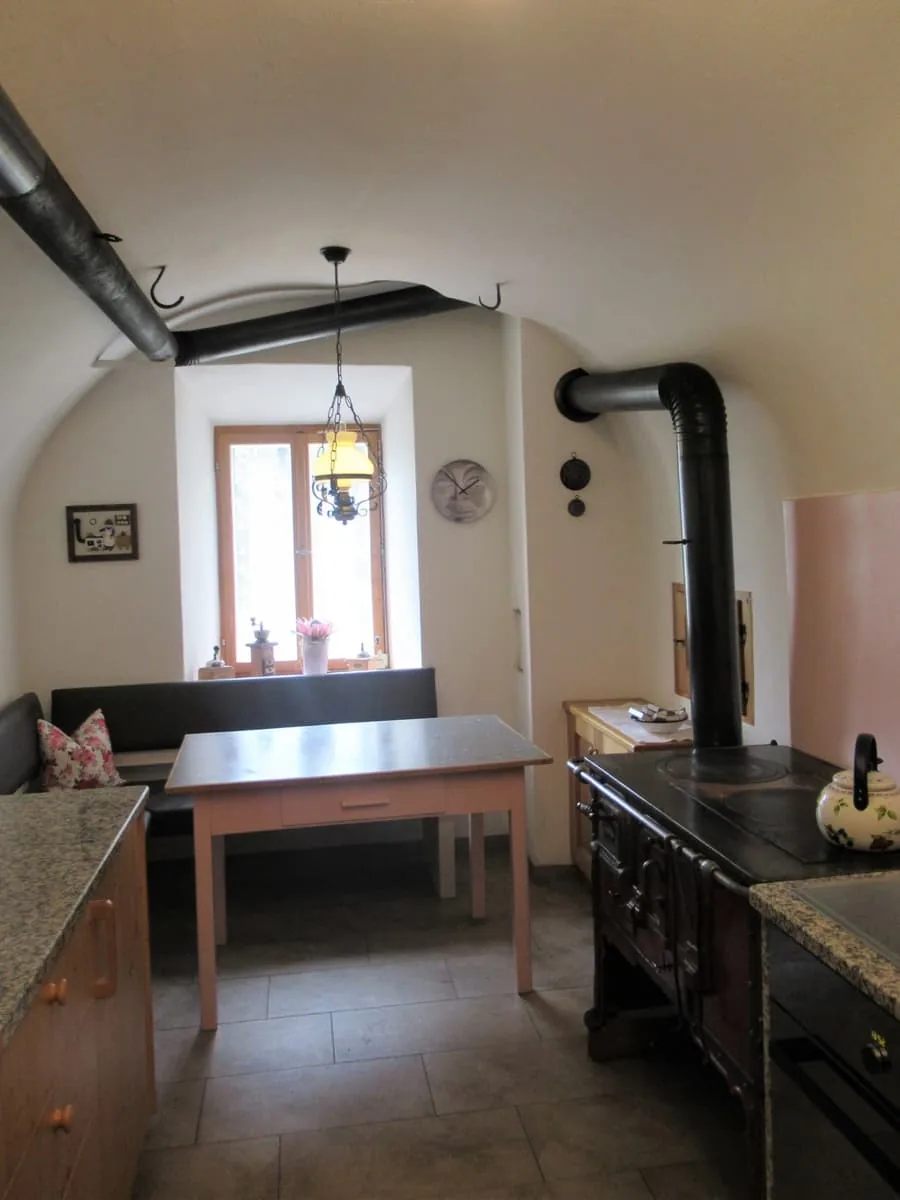Küche mit gewölbter Decke und tief eingesetztem Fenster, rechts der schwarze Holzherd, davor ein Tisch mit Sitzbank.