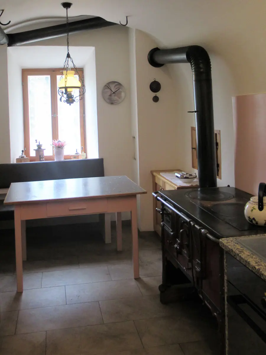 Küche mit gewölbter Decke: das tief eingesetzte Fenster in der weiss verputzten Laibung, davor Sitzbank und Tisch, rechts der schwarze Holzherd mit dem Ofenrohr.