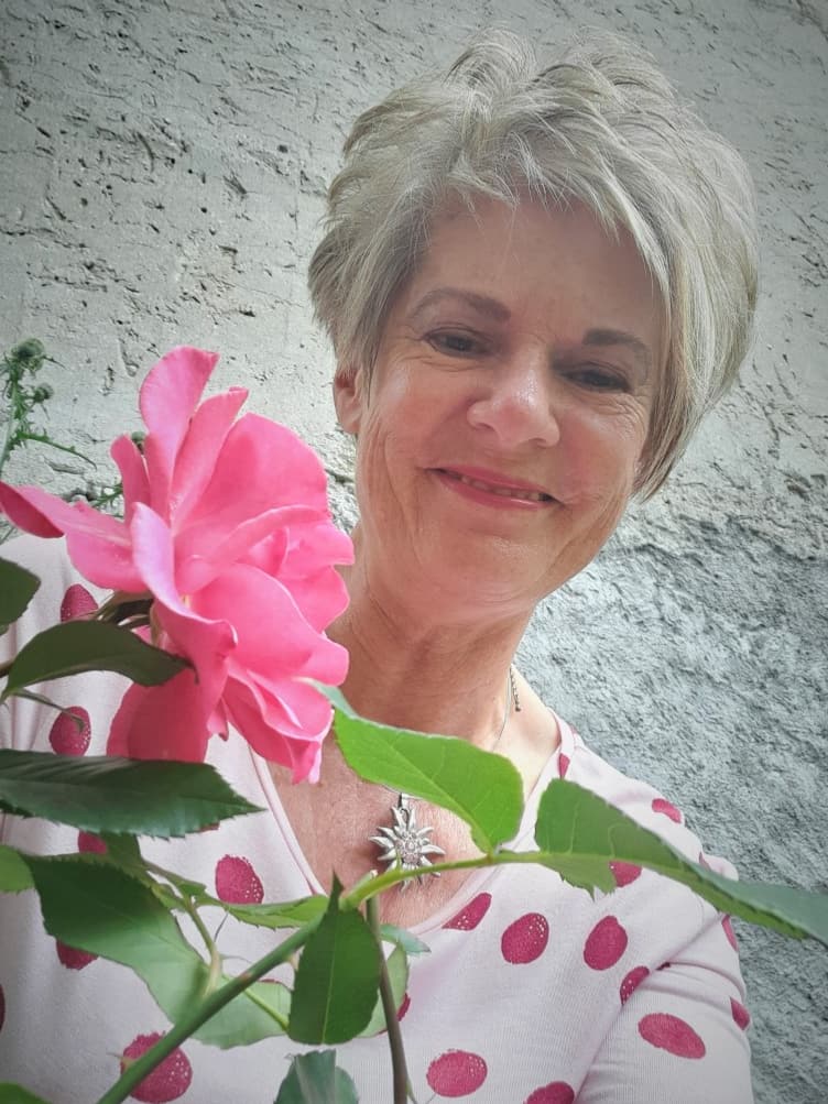 Marina Margadant im Garten neben einer rosa Rose vor der verputzten Wand ihres Hauses.
