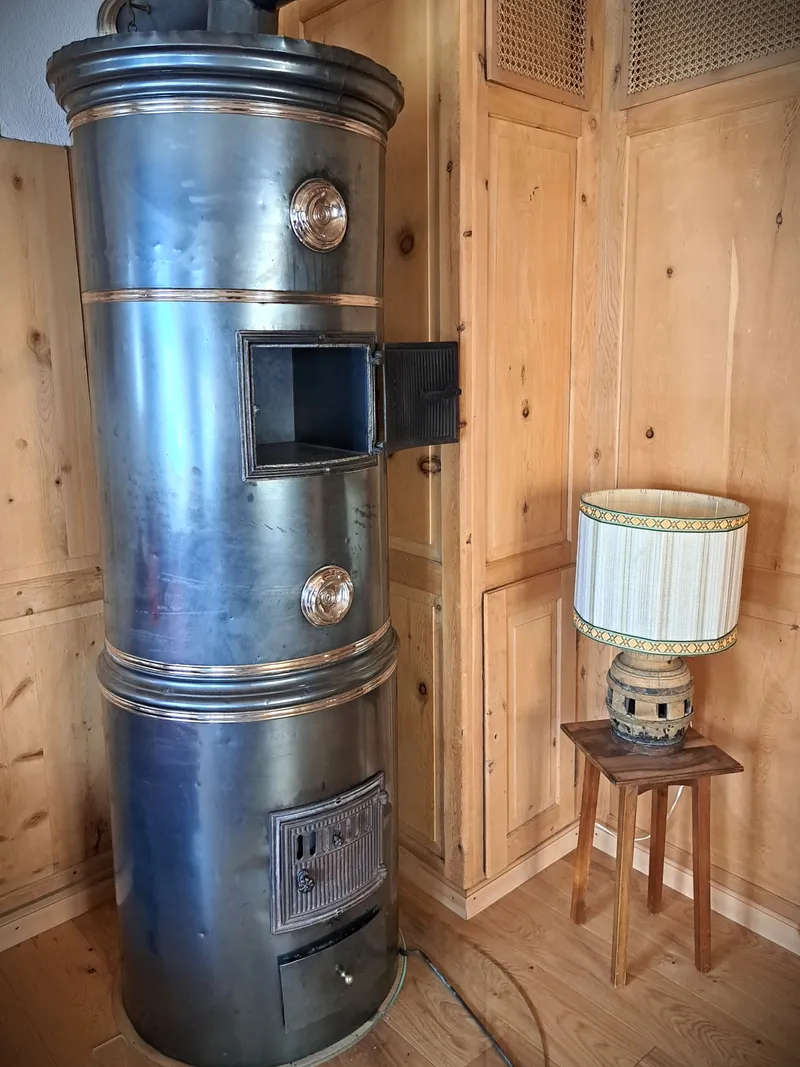 Hoher Ofen aus Metall mit Kupferringen in einer Stube mit hellen Holzwänden, daneben ein Hocker mit Lampe.