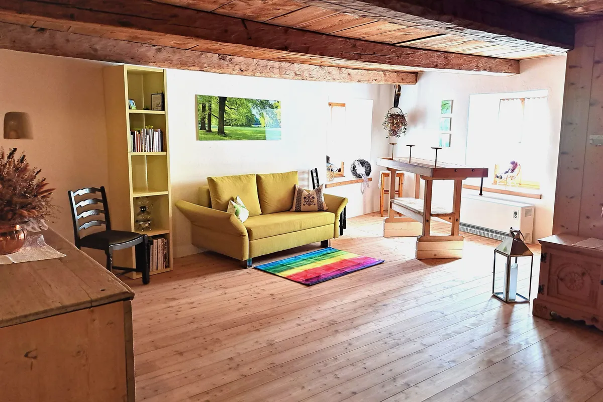 Heller Raum mit Holzbalken und Dielenboden, gelbes Sofa, Stehpult und Fenster, Ort für die Seminare.