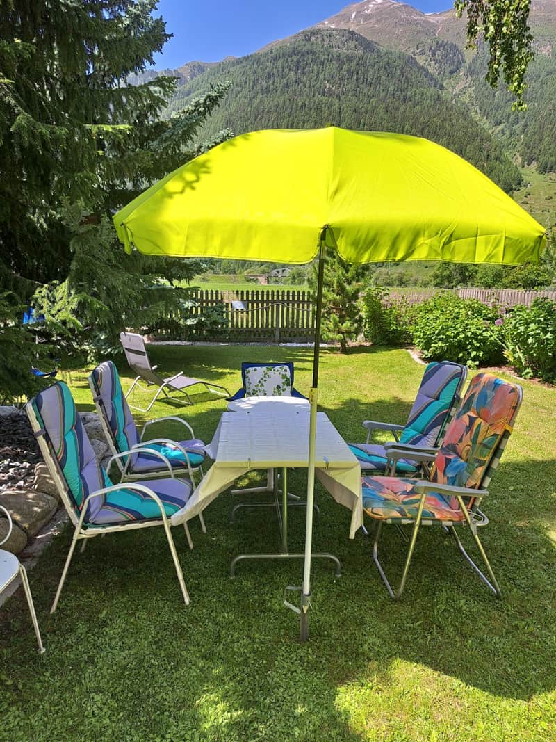 Gartenstühle und Tisch unter einem gelben Sonnenschirm auf dem Rasen, dahinter die Wiesen und Berge des Engadins.