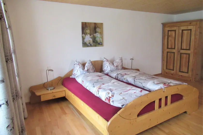 Gästezimmer mit einem Doppelbett aus hellem Holz, rotem Leintuch und geblümter Bettwäsche, rechts ein alter Holzschrank.