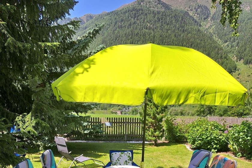 Gelber Sonnenschirm über Gartenstühlen auf dem Rasen, hinter dem Holzzaun bewaldete Hänge und die Berge des Engadins.