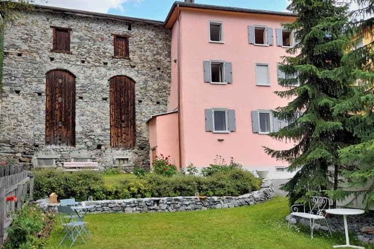 Das rosa verputzte Haus mit grauen Fensterläden neben dem alten Steinstall mit zwei hohen Holztoren, davor der Garten mit Rasen und Gartentisch.