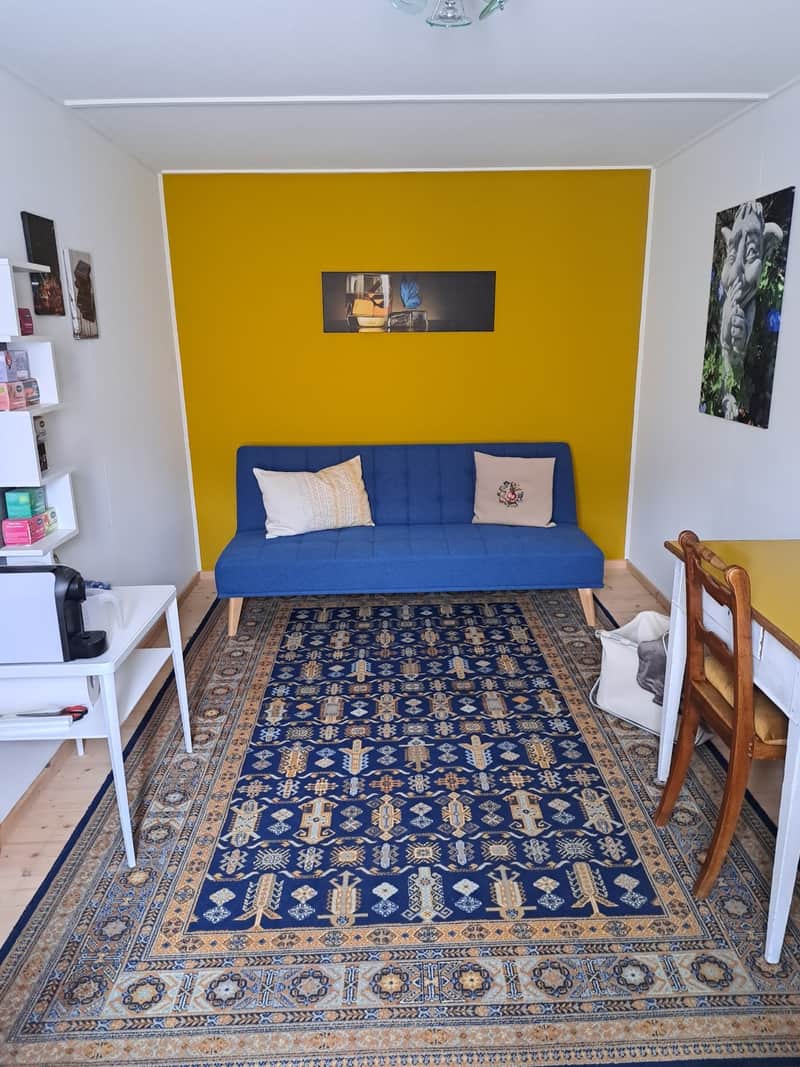 Stüva mit ockergelber Wand, blauem Sofa, gemustertem Teppich und Esstisch.