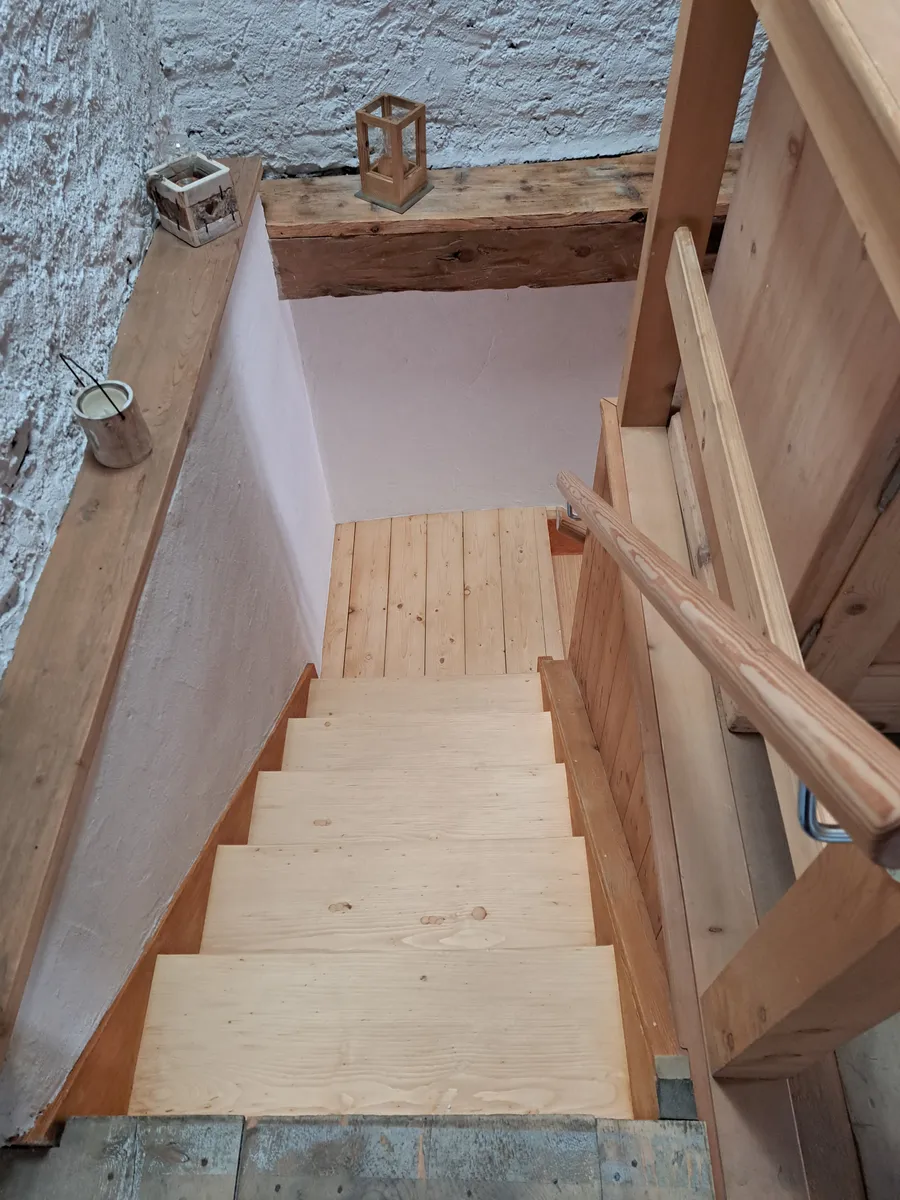 Blick in das Treppenhaus: helle Holzstufen mit Handlauf, oben auf dem Balken eine Laterne.